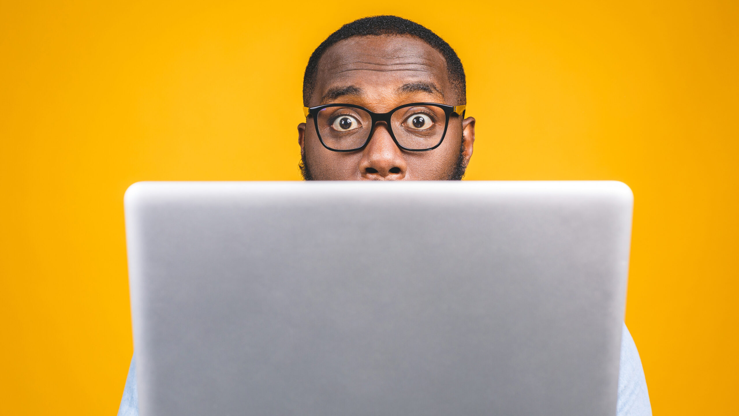 Young surprised african american man standing and using laptop computer isolated over yellow background.